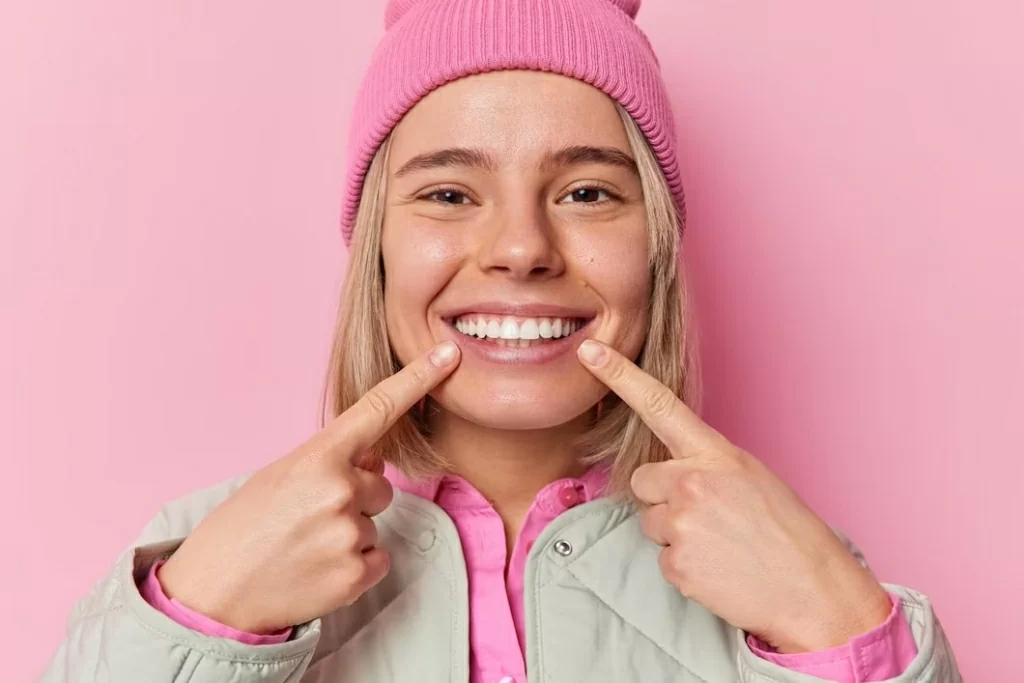 cute-positive-caucasian-woman-points-index-fingers-toothy-smile-shows-her-perfect-teeth-wears-hat-jacket-being-good-mood-isolated-pink-background-people-happiness-concept.jpg