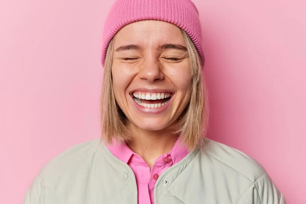 positive-authentic-european-woman-smiles-broadly-shows-white-teeth-giggles-happily-with-closed-eyes-wears-hat-jacket-poses-studio-against-pink-background-people-emotions-concept_273609-59737.jpg